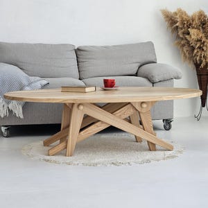 May include: Oval wooden coffee table with a distinctive, geometric base design. The light-coloured wood table is in a living room setting with a grey sofa, a book, and a red cup. The table is approximately 140 cm long.
