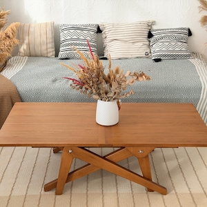 May include: A wooden coffee table with a natural finish, featuring a folding design. The table is set on a striped rug, with a vase of dried flowers on top. Pillows and a blanket are on the sofa in the background.