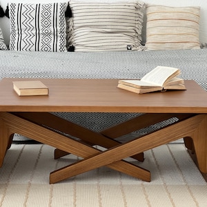 K&ouml;nnte beinhalten: Ein rechteckiger Couchtisch aus Holz mit glatter brauner Oberfl&auml;che und einem markanten X-f&ouml;rmigen Untergestell. Die Tischplatte ist flach und weist eine nat&uuml;rliche Holzmaserung auf, die eine stabile Ablagefl&auml;che f&uuml;r B&uuml;cher bietet.