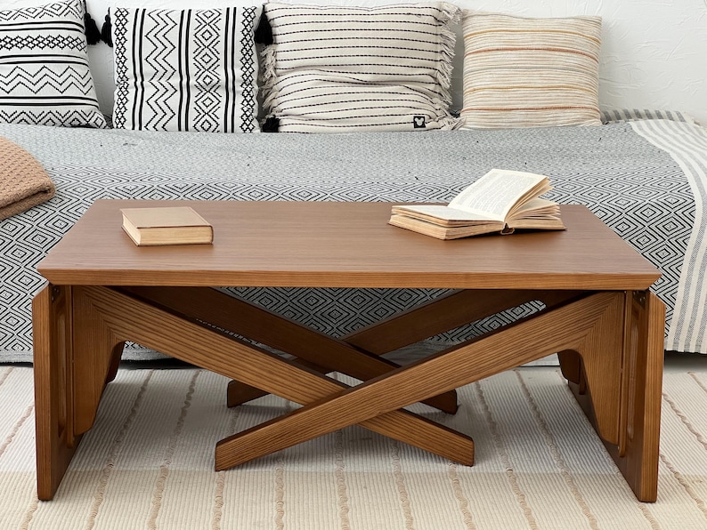 K&ouml;nnte beinhalten: Ein rechteckiger Couchtisch aus Holz mit glatter brauner Oberfl&auml;che und einem markanten X-f&ouml;rmigen Untergestell. Die Tischplatte ist flach und weist eine nat&uuml;rliche Holzmaserung auf, die eine stabile Ablagefl&auml;che f&uuml;r B&uuml;cher bietet.