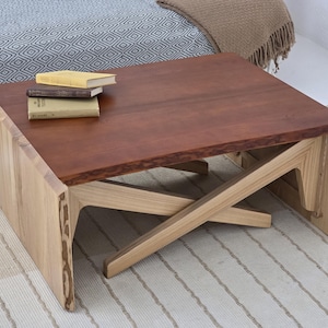 Peut inclure: Une table basse en bois faite à la main, avec un plateau brun rougeâtre riche et des pieds en bois de couleur claire. La table est de forme carrée et mesure environ 91 cm de long et de large. Trois livres sont empilés dessus.