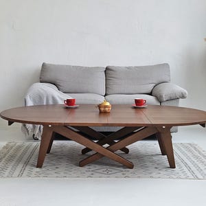 May include: A wooden oval coffee table with a dark brown finish, featuring a unique cross-legged base. Two red cups and a teapot sit on the table. The table is placed on a patterned rug in front of a gray sofa.