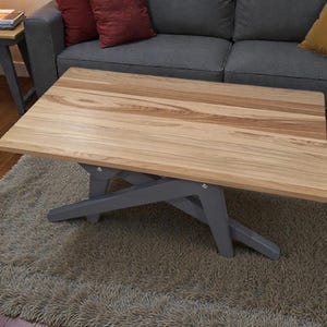 Puede incluir: Mesa de centro rectangular de madera clara con patas grises. La mesa está sobre una alfombra beige. Un sofá gris con cojines rojos y amarillos se encuentra en el fondo.