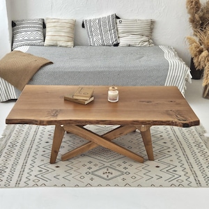 May include: A rectangular wooden coffee table with a natural edge, supported by a crossed-leg base. The table is placed on a patterned rug. Pillows and a blanket are on a bed in the background. A candle and books are on the table.