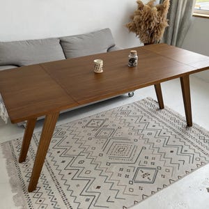 K&ouml;nnte beinhalten: Ein rechteckiger Esstisch aus Holz mit glatter, dunkelbrauner Oberfl&auml;che und vier schr&auml;gen Beinen. Die Tischplatte weist eine sichtbare Naht auf, die auf ein ausziehbares Design hindeutet. Zwei kleine Dekoartikel stehen in der Mitte.