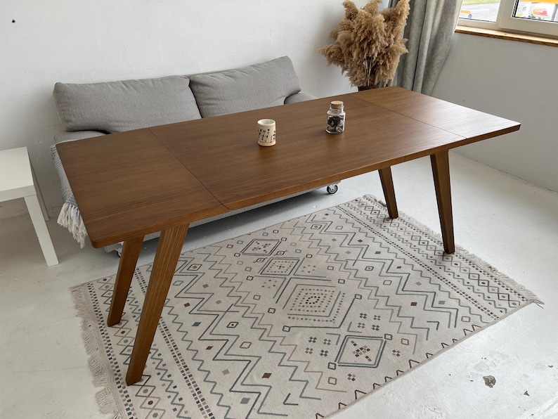 K&ouml;nnte beinhalten: Ein rechteckiger Esstisch aus Holz mit glatter, dunkelbrauner Oberfl&auml;che und vier schr&auml;gen Beinen. Die Tischplatte weist eine sichtbare Naht auf, die auf ein ausziehbares Design hindeutet. Zwei kleine Dekoartikel stehen in der Mitte.