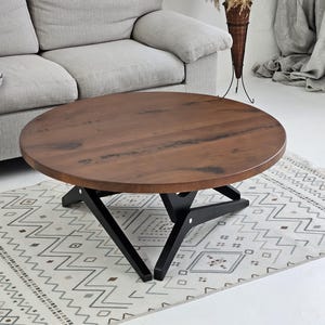 May include: A round, dark brown wooden coffee table with a black, geometric base. The table sits on a patterned rug in a living room setting, near a gray sofa. The table's diameter is approximately 36 inches.