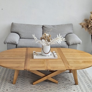 May include: A light wood oval coffee table with a cross-shaped base. The table has two leaves that can be extended to make it larger. The table is sitting on a white and grey patterned rug in front of a grey sofa.