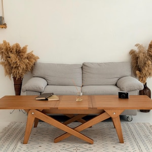 May include: A wooden coffee table with an expandable top, set in a living room. The table is a light brown color and has a unique leg design. A gray sofa is behind the table, and a rug is beneath it. Decorative dried plants are in vases on either side.