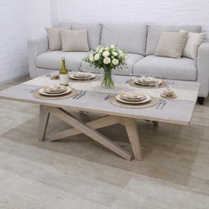 Könnte beinhalten: Ein hellgrauer rechteckiger Couchtisch mit Holzfuß, gedeckt für eine Mahlzeit. Der Tisch steht vor einem hellgrauen Sofa mit beigen Kissen. Eine Vase mit weißen Blumen, eine Weinflasche, Teller und Besteck stehen auf dem Tisch.