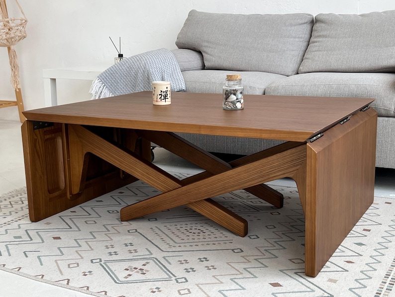K&ouml;nnte beinhalten: Ein rechteckiger Couchtisch aus Holz mit einem satten braunen Finish und sichtbarer Holzmaserung. Der Tisch verf&uuml;gt &uuml;ber ein einzigartiges X-f&ouml;rmiges Basisdesign mit klappbaren Seitenteilen, die durch Metallscharniere verbunden sind. Ein kleines wei&szlig;es Gef&auml;&szlig; mit schwarzem Text steht darauf.