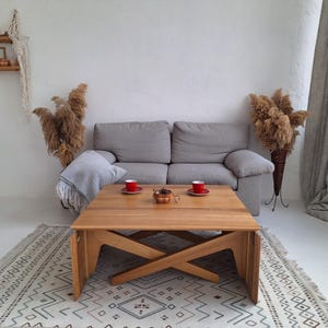 Può includere: Scena di soggiorno con un divano grigio chiaro, un tavolino in legno e un tappeto fantasia. Due tazze rosse sul tavolo. Piante secche e una mensola decorativa completano l'arredamento.