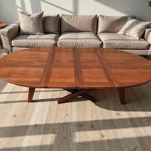 Può includere: Tavolino ovale in legno con una ricca finitura marrone. Il tavolo presenta un design unico con un supporto centrale a forma di croce e un piano del tavolo a pannelli multipli. Il tavolo è in un soggiorno con un divano grigio e cuscini fantasia.