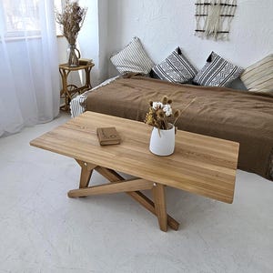 May include: A light brown wooden folding table with a rectangular top and angled legs. A small, closed book rests on the table. A white vase with dried flowers sits on the table. The table is in a room with a bed and pillows.