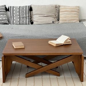 May include: A rectangular wooden coffee table with a smooth brown finish and a cross-legged base design. The table features a flat top surface with a closed book on the left side and an open book on the right side, showcasing a natural wood grain.