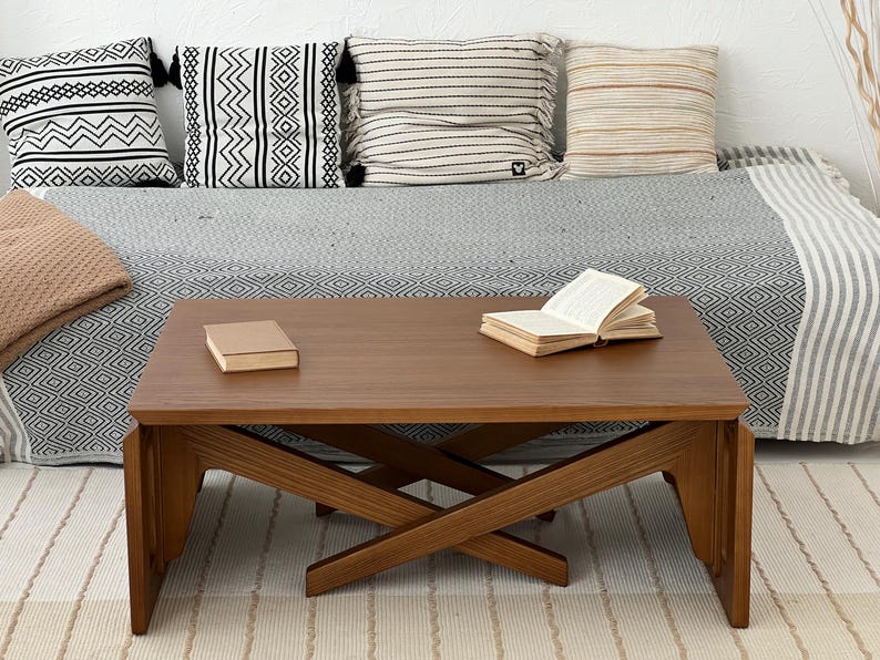 K&ouml;nnte beinhalten: Ein rechteckiger Couchtisch aus Holz mit glatter brauner Oberfl&auml;che und einem kreuzf&ouml;rmigen Untergestell. Der Tisch hat eine flache Oberseite, auf der links ein geschlossenes Buch und rechts ein aufgeschlagenes Buch liegen, mit sichtbarer Holzmaserung.