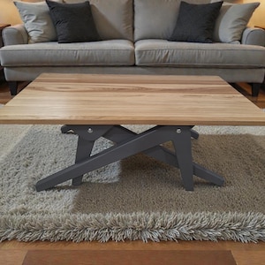 May include: A square wooden coffee table with a light brown top and gray legs. The table sits on a shaggy, light brown rug. The table is in front of a gray sofa with black pillows.
