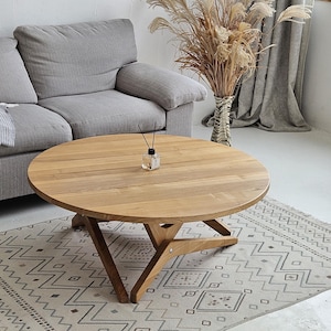 Può includere: Un tavolino da caffè rotondo in legno con finitura in legno naturale e base geometrica. Il tavolino è posizionato su un tappeto a motivi bianchi e grigi in un soggiorno.