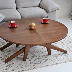 May include: A circular, wooden coffee table with a dark brown finish. The table has a distinctive leg design and is placed on a patterned rug. Two red cups and a small basket are on the table. A grey sofa is in the background.
