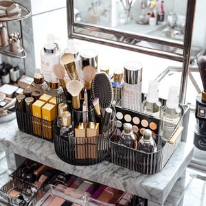 Puede incluir: Un tocador con varios productos y accesorios de belleza. Organizadores transparentes y negros contienen brochas de maquillaje, botellas y paletas. La disposición incluye productos para el cuidado de la piel, perfumes y un espejo, todo sobre una superficie de mármol.