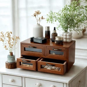 Puede incluir: Un pequeño mueble de cajones de madera oscura con cuatro cajones, dos abiertos. Los cajones tienen frentes de vidrio y tiradores de metal. Varias botellas de cosméticos y jarrones con flores están encima del mueble.