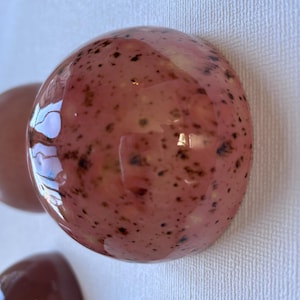 May include: A glossy, spherical ceramic object with a light pink base colour and dark brown speckles distributed across its surface. The object has a smooth, reflective finish that catches light, creating bright highlights on its rounded top.