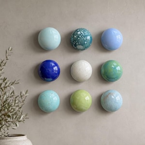 May include: A collection of nine circular, smooth, and glossy ceramic wall decorations arranged in a three-by-three grid. The spheres feature various finishes, including solid light blue, deep cobalt, speckled white, mottled green, and textured teal.