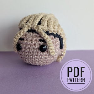 Puede incluir: Una pequeña cabeza amigurumi de ganchillo, redonda, con cabello de hilo beige claro, una diadema negra, cara rosa pálido y dos ojos de seguridad de plástico negro. La figura tiene un pequeño pendiente de aro plateado. Texto: PDF PATTERN.