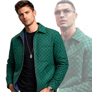 Op de afbeelding: Groene jas met een herhalend zwart patroon en een ritssluiting. De jas wordt gedragen over een zwart shirt en een zilveren ketting met een hondenpenning hanger. Een horloge is zichtbaar op de pols.