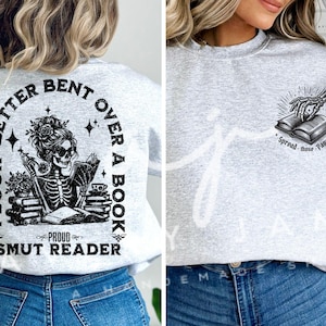 May include: Grey sweatshirt with a black and white graphic of a skeleton reading a book. The text on the graphic reads "I look better bent over a book, Proud Smut Reader".