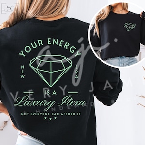 May include: Black sweatshirt with a diamond graphic and the text "YOUR ENERGY NEW ERA IS A LUXURY ITEM NOT EVERYONE CAN AFFORD IT". A smaller diamond graphic is on the upper right. The design is in a light green color.