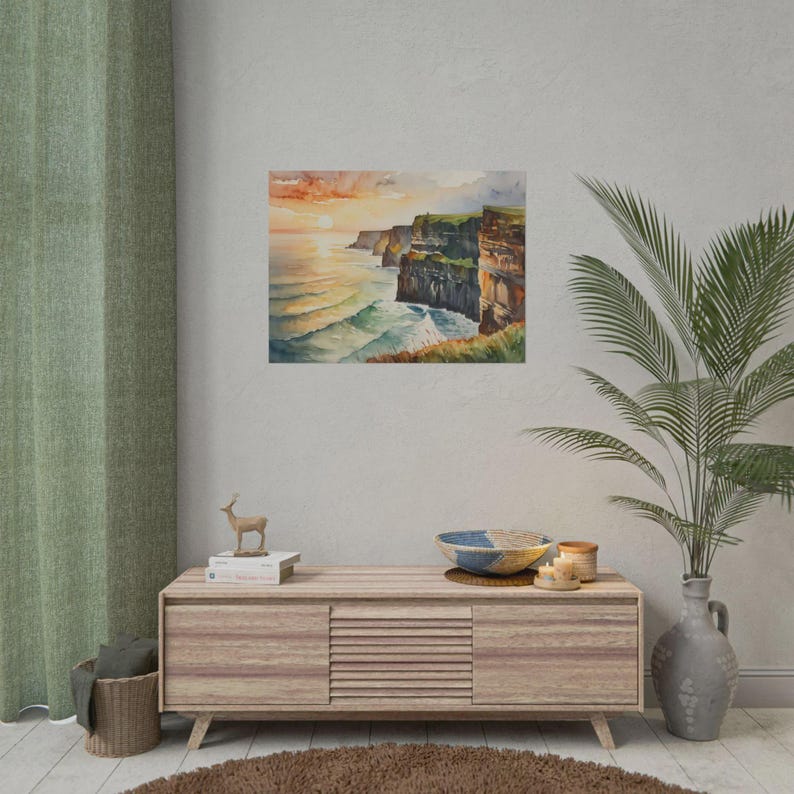 May include: Watercolor print of a coastal landscape featuring cliffs, ocean waves, and a sunset. The artwork is displayed on a wall, with a wooden cabinet and decorative items in the foreground. The color palette includes shades of orange, blue, and green.