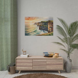 May include: Watercolor painting of a coastal landscape featuring cliffs and ocean waves at sunset. The artwork depicts a vibrant sky with orange and yellow hues, reflecting on the water. The cliffs are brown and green, with white waves crashing below.