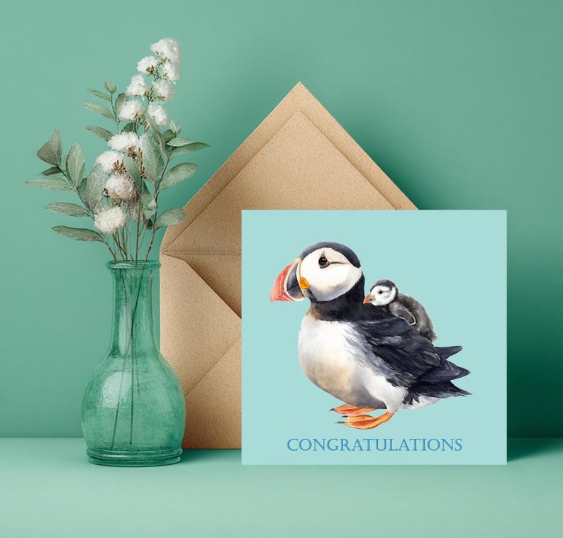 Puede incluir: Una tarjeta de felicitaci&oacute;n con una ilustraci&oacute;n en acuarela de un frailecillo que lleva un beb&eacute; frailecillo en la espalda. La tarjeta tiene un fondo azul claro y la palabra "FELICIDADES" en azul. Un jarr&oacute;n de cristal verde con flores blancas a la izquierda.