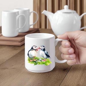 Puede incluir: Taza de cer&aacute;mica blanca con una ilustraci&oacute;n de acuarela de dos frailecillos sobre hierba verde. La taza tiene un asa curva y est&aacute; sujeta por una mano. Otras tazas blancas y una tetera est&aacute;n en el fondo.