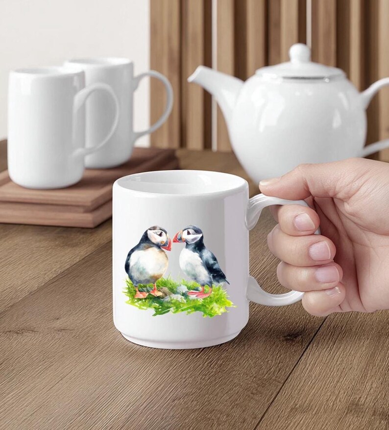 Puede incluir: Taza de cer&aacute;mica blanca con una ilustraci&oacute;n de acuarela de dos frailecillos sobre hierba verde. La taza tiene un asa curva y est&aacute; sujeta por una mano. Otras tazas blancas y una tetera est&aacute;n en el fondo.