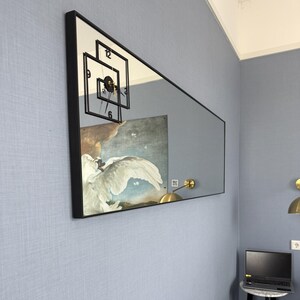 May include: A large rectangular mirror with a black frame hangs on a light blue wall. The mirror reflects a clock with black hands and Roman numerals, and a painting of a white bird with a blue sky background.