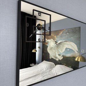 May include: A black framed rectangular mirror with a clock face reflected in the glass. The clock face is made of black squares with white numbers and hands. The mirror reflects a bedroom with a white bed, black curtains, and a painting of a white bird on a blue background.