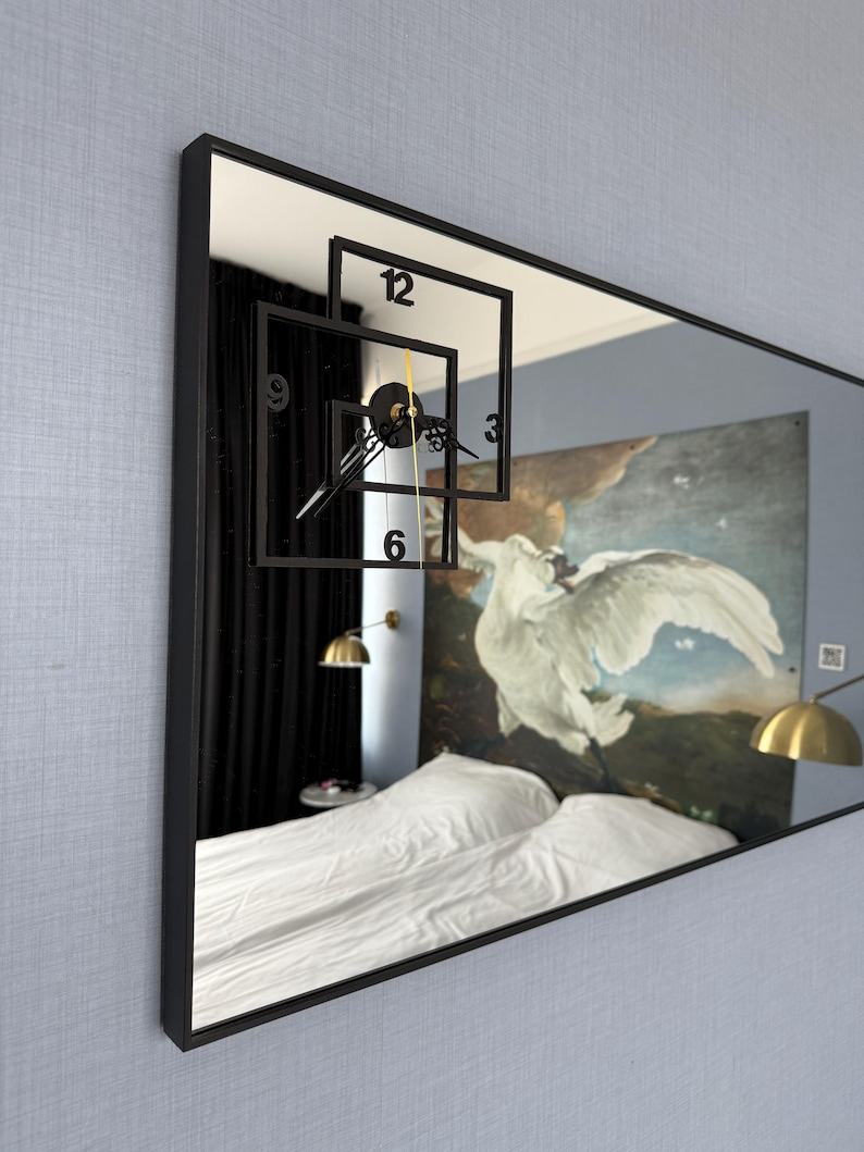 May include: A black framed rectangular mirror with a clock face reflected in the glass. The clock face is made of black squares with white numbers and hands. The mirror reflects a bedroom with a white bed, black curtains, and a painting of a white bird on a blue background.