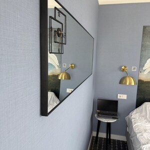 May include: A large rectangular mirror with a black frame hangs on a light blue wall. The mirror reflects a clock with black Roman numerals and a gold lamp. The mirror is positioned in a corner of the room.