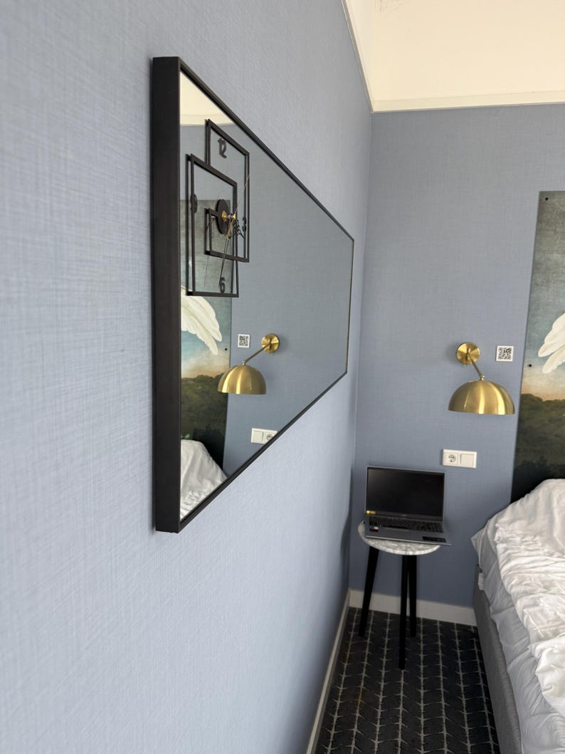 May include: A large rectangular mirror with a black frame hangs on a light blue wall. The mirror reflects a clock with black Roman numerals and a gold lamp. The mirror is positioned in a corner of the room.