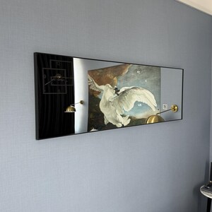 May include: A black framed rectangular mirror with a white swan painting reflected in it. The swan is flying with its wings spread wide. The painting is set against a blue sky with white clouds. There are two gold wall sconces on either side of the mirror.