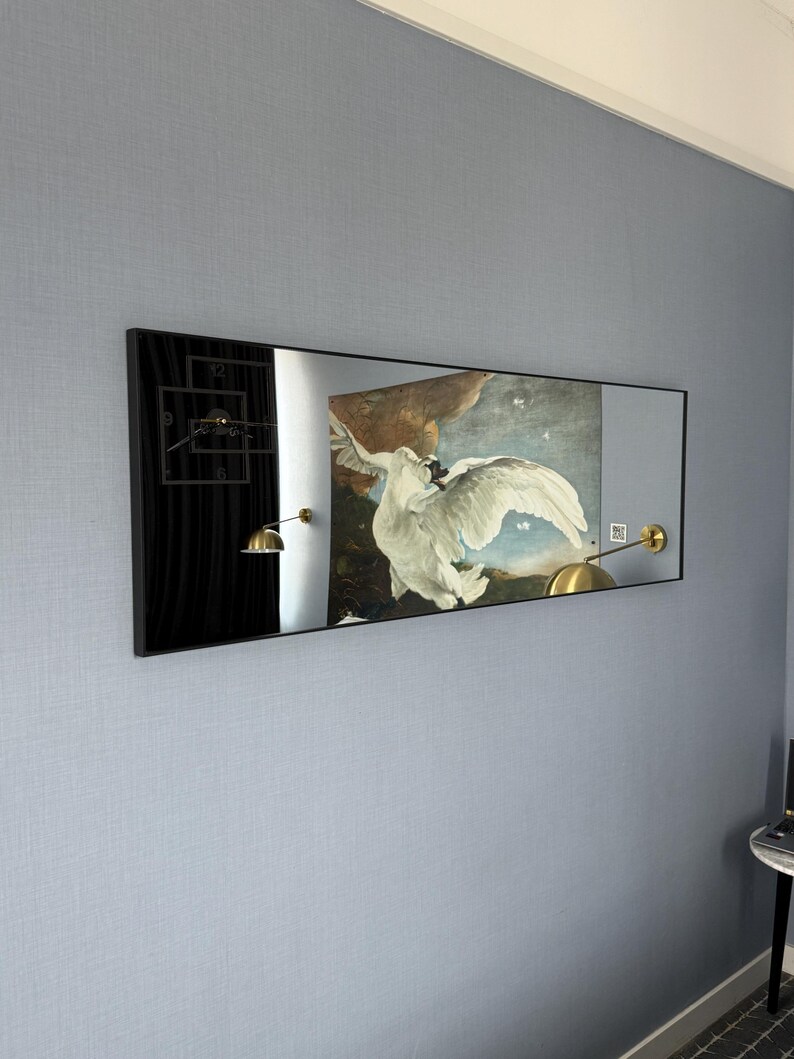 May include: A black framed rectangular mirror with a white swan painting reflected in it. The swan is flying with its wings spread wide. The painting is set against a blue sky with white clouds. There are two gold wall sconces on either side of the mirror.