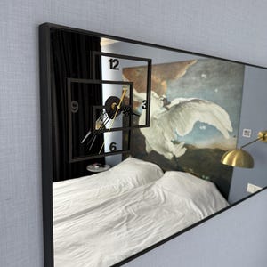 May include: A black framed mirror with a clock face reflected in the glass. The clock face is made of black numbers and hands on a white background. The mirror is hanging on a light blue wall.