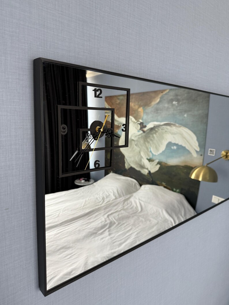May include: A black framed mirror with a clock face reflected in the glass. The clock face is made of black numbers and hands on a white background. The mirror is hanging on a light blue wall.