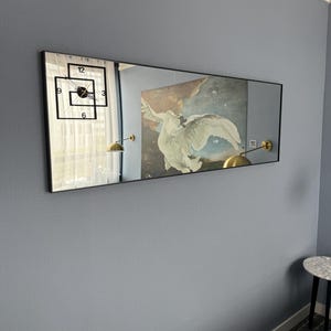 May include: A long rectangular mirror with a black frame hangs on a light blue wall. The mirror reflects a clock with black hands and numbers, a white curtain, and a painting of a white bird with a brown background. Two gold wall lamps are visible in the reflection.