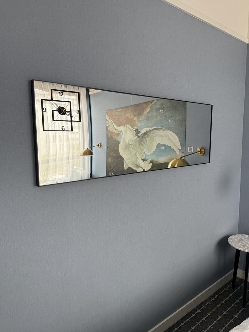 May include: A long rectangular mirror with a black frame hangs on a light blue wall. The mirror reflects a clock with black hands and numbers, a white curtain, and a painting of a white bird with a brown background. Two gold wall lamps are visible in the reflection.