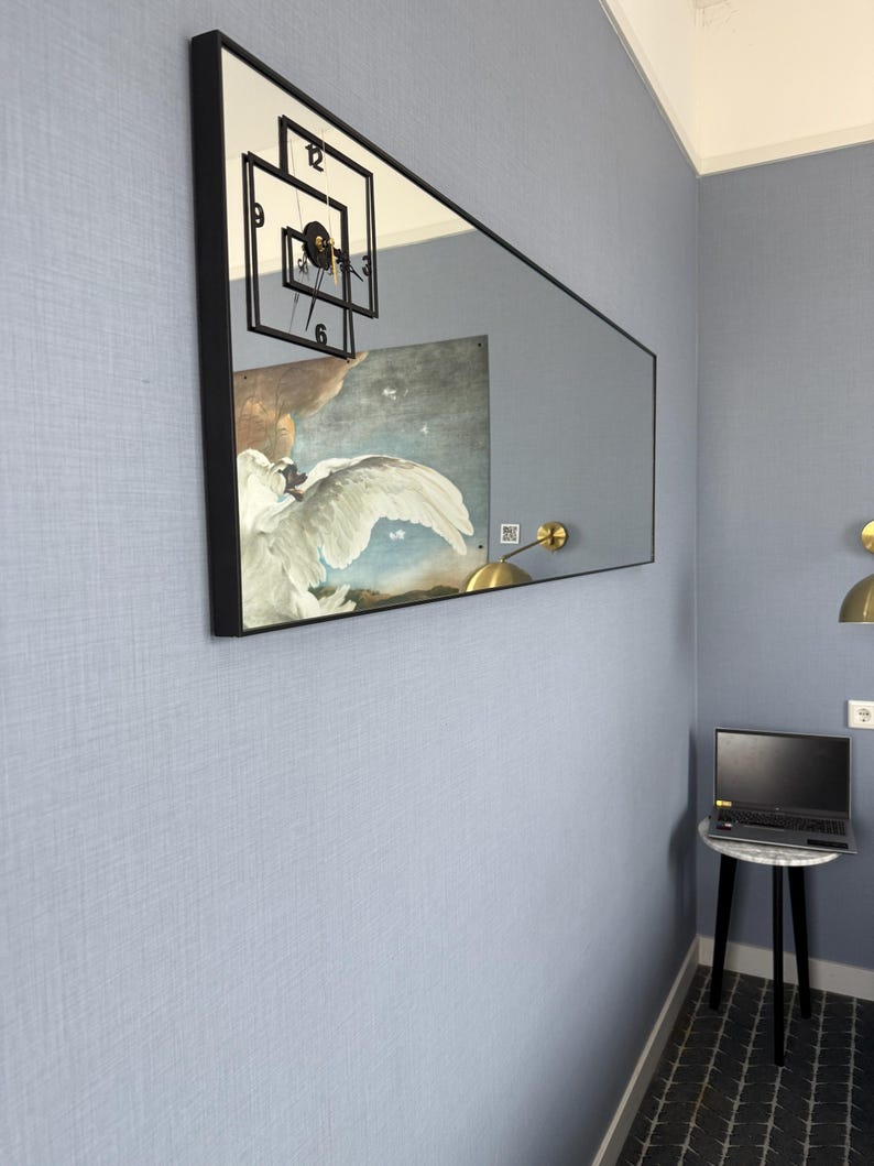 May include: A large rectangular mirror with a black frame hangs on a light blue wall. The mirror reflects a clock with black hands and Roman numerals, and a painting of a white bird with a blue sky background.