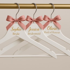 Puede incluir: Tres perchas de madera blancas con lazos de satén rosa y letras doradas. Las perchas dicen "Sophie Novia", "Jessica Dama de honor" y "Olivia Dama de honor".