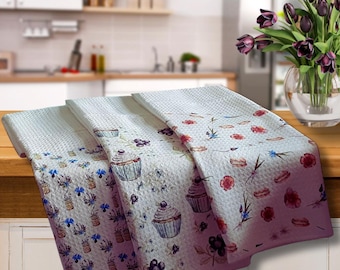 Könnte beinhalten: Drei weiße Küchentücher mit Waffelstruktur. Jedes Tuch hat ein anderes Design aus Blumen und Desserts, darunter Cupcakes, Blumen und Beeren. Die Tücher werden auf einer Holzoberfläche präsentiert.